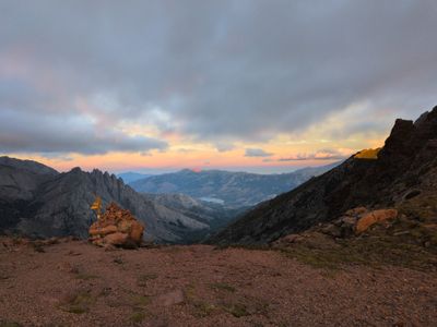 Photo of a mountain col at sunset. Cairn with waymark signs on the left. Rocky slopes and a green valley in the center