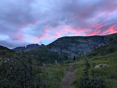 A photo. Mountain cliff in the background, small bushes in the foreground. A distant person with a backpack in the middle. Pink sunset clouds above the cliff.